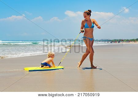 Happy family having fun - mother run and pull baby son on surfing board by water pool along sea surf of beach. Active parents and kids lifestyle people activity on summer beach holiday with children