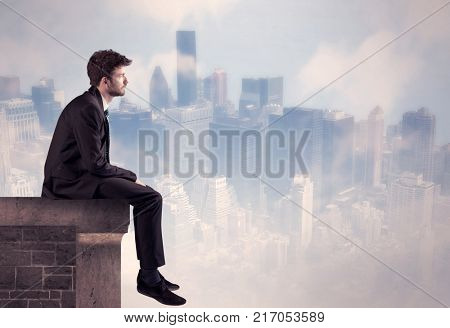 A young businessman sitting at the edge of a building in front of a city center scape background with tall buildings and clouds concept