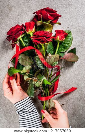 Celebratory background Valentine's day. The girl (hands in the picture) makes a bouquet of red roses tie with a red ribbon. On a gray stone table top view copy space
