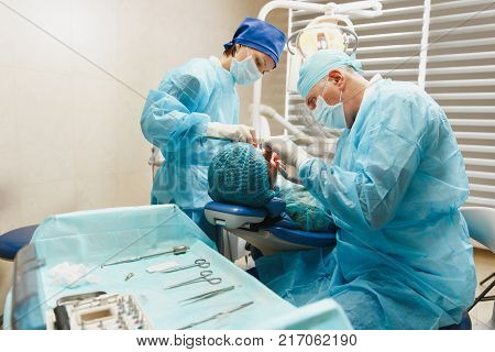 Male Old Professional Dentist Surgeon In Uniform And Female Assistant Helps To Performing Operation