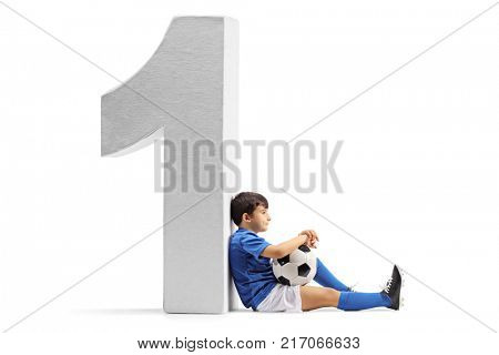 Pensive little footballer sitting on the floor and leaning against a cardboard number one isolated on white background