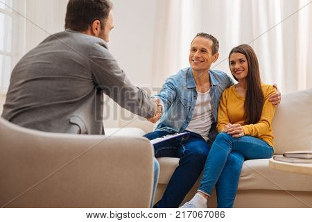 See you tomorrow. Positive appealing kind couple saying goodbye to a psychologist and sitting on the sofa while man shaking doctors hand