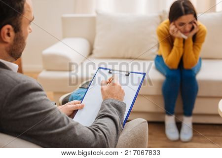 Professional commentary. Male psychologist writing down commentary while depressed female patient sitting on the sofa and looking aside