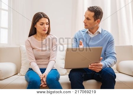 Great plan. Appealing concerned man putting laptop on his knee while demonstrating his plan to depressed  wife while she sitting