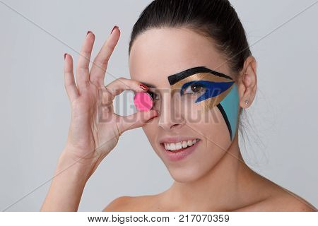 Portrait of a brunette with a bunch on her head, a woman with creative geometric make-up on the face, black lines, holding a pink paint for make-up, near the eye, smiling, looking at the camera