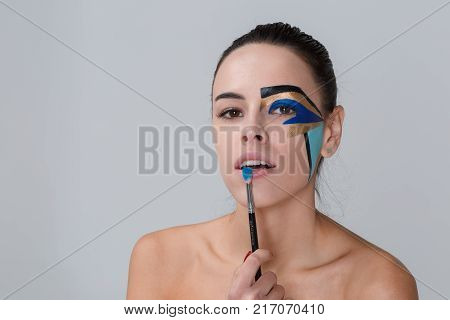 Portrait of a brunette with a bunch on her head, a woman with creative geometric make-up on the face, black lines, holding a make-up brush near her lips, looks into the camera, her teeth are seen.