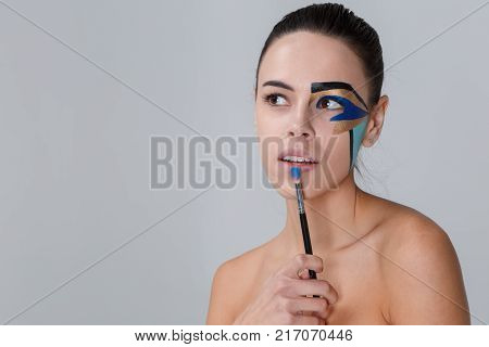 Portrait of a brunette with a bunch on her head, a woman with creative geometric make-up on the face, black lines, holding a make-up brush near her lips, looking away, you can see teeth.