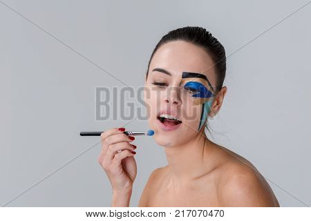 Portrait of a brunette with a bunch on her head, a woman with creative geometric make-up on the face, black lines, holding a make-up brush near the chin, looking down, opening her mouth