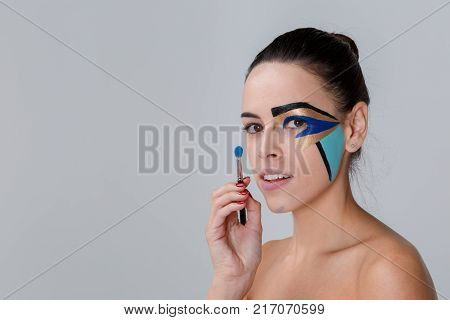 Portrait of a brunette with a bunch on her head, a woman with creative geometric make-up on the face, black lines, holding a make-up brush near her face, looks into the camera, her mouth slightly open