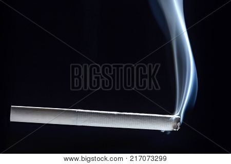 A completely white cigarette begins to smolder and smokes against a dark background.