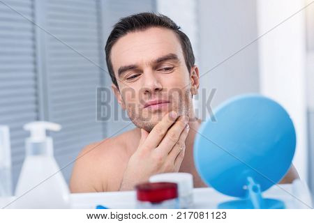 Great profile. Attentive good looking young man observing his face while touching his chin and looking at the mirror