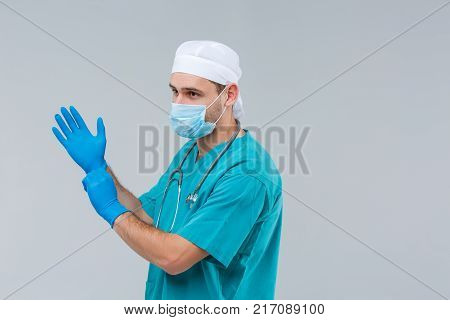 Close-up of a doctor in uniform, in profile, wearing a mask on a gray background, a man in his arms dresses blue, rubber gloves, on his neck hangs strobo, looks away.