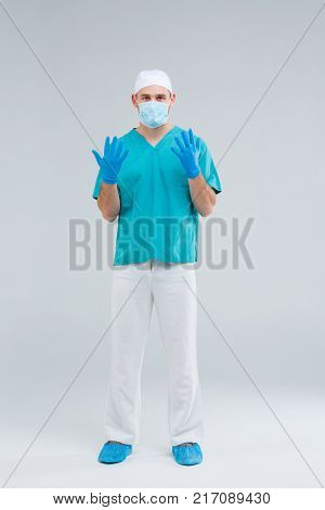 A doctor in uniform, full-length, front view, on a gray background, a beanie on his head, on the legs of a shoe cover, in a bandage, in blue gloves, spread his fingers, looks into the camera.