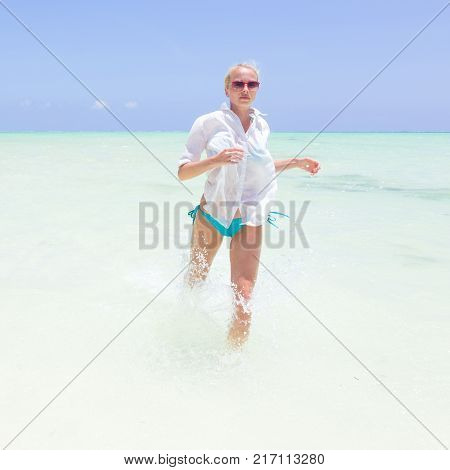 Young slim fit woman wearing white beach tunic running in sea water making water splashes with her legs. Vacation concept. Summer mood. Tropical beach setting. Paje, Zanzibar, Tanzania.