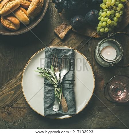 Fall holiday table decoration setting. Plate with linen napkin, fork and spoon, candle, fresh fruits on board, bread buns, olive tree branch over wooden background, top view, copy space, square crop