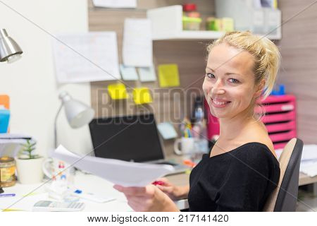 Business and entrepreneurship consept. Beautiful blonde business woman talking on mobile phone in colorful modern creative working environment accepting papers. Female multi tasking.