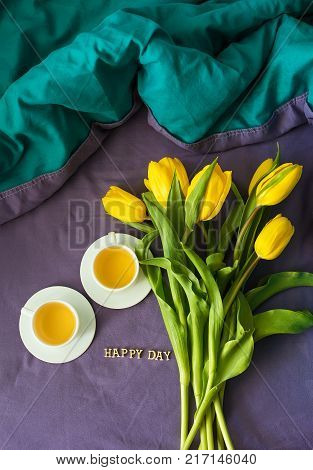 Yellow tulips and two cups of tea in bed, close-up, lettering happy day