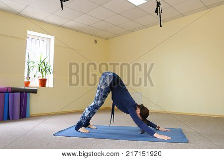 Sportswoman focused on stretch before Pilates class. Girl doing warm up for legs on mat. Female has hair pulled back, concentrated look. Athlete dressed in dark top, yoga legging trains in fitness room. Concept of online training, customizable workouts.