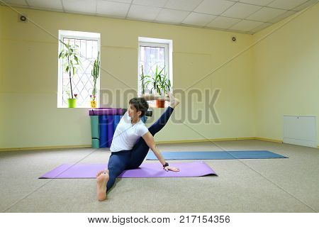 Sportswoman focused on stretch before Pilates class. Girl doing warm up for legs on mat. Female has hair pulled back, concentrated look. Athlete dressed in dark top, yoga legging trains in fitness room. Concept of online training, customizable workouts.