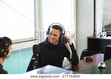 Cute female radio host and funny guy with headphones and talking are direct inclusion of radio, sitting in chair in radio studio. In office ponoramic large windows on desktop are microphones and mixing consoles, broadcast processor, special devices 