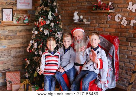 Merry brothers boys sitting on Santa Clauss lap and waving their palms camera rejoice. Claus with gray long beard dressed in red suit white trim pointed cap pompon end. Charming men of European appearance warm sweaters blue jeans socks, sit large soft arm
