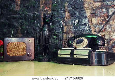 The figure of a black cat looking next to the old telephone and radio on the table.