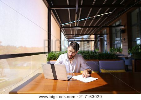 Inventory strategist solving problems and using laptop at cafe table. Clever hardworking man dressed in white shirt working with papers. Concept of  analyzing model forecast  to design contingency plans and catalog displays to make successful 