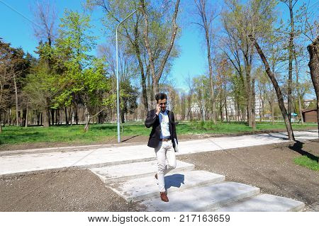 Guy walking in park and smiling, slow motion. Man dressed in blue shirt, white jeans and brown shoes. Boy has black hair and beard. Concept of places for rest and clean parks and cheeps fashion clothes for man