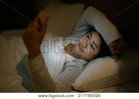 Young asian woman on bed late at night texting using mobile phone sleepy and tired in internet communication overuse and smartphone addiction. Wife using smartphone waiting husband with boring emotion