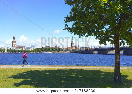 The girl in sports clothes runs along the embankment of the Daugava River in Riga against the backdrop of the old city in the summer