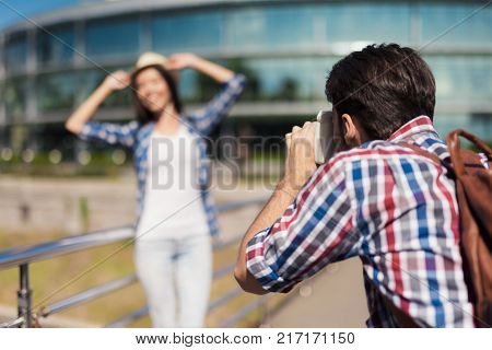 A guy takes a picture of a beautiful girl against a beautiful modern building. The girl is happy to be posing and smiling. She has a white hat on her head.
