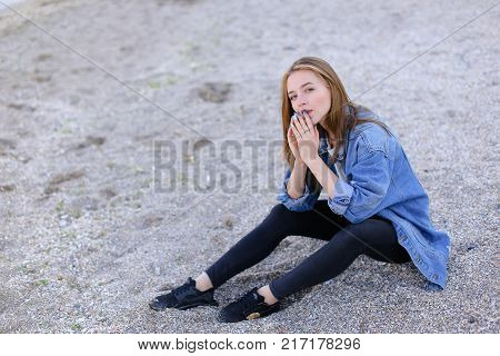 Dreamy girl relaxes and enjoys rest on seaside, throws head back and shakes hair, sitting on sandy beach on Black Sea shore on clear evening. Young European-looking woman with blond mid-length hair dressed in white jacket over which blue denim jacket,