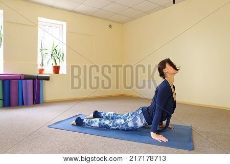 Sportswoman focused on stretch before Pilates class. Girl doing warm up for legs on mat. Female has hair pulled back, concentrated look. Athlete dressed in dark top, yoga legging trains in fitness room. Concept of online training, customizable workouts.
