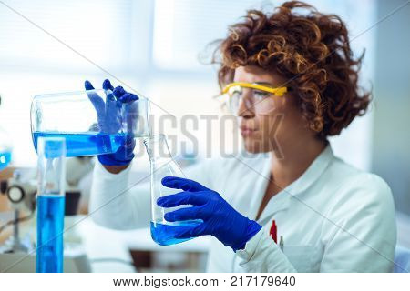 Young attractive female scientist in protective eyeglasses and gloves using test tube with blue liquid sample substance probe in the scientific chemical research laboratory