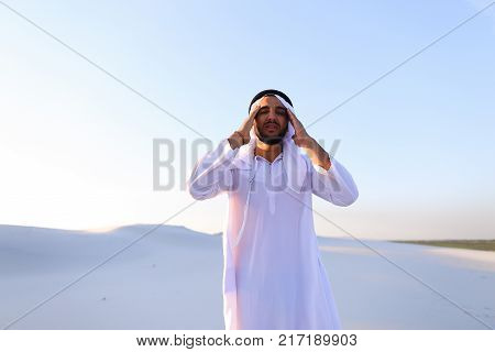 Portrait of tired Arab man, who holds head and feels migraine, tries to calm pain and massages whiskey, standing in middle of bottomless desert with snow-white sand on sunny summer day. Swarthy Muslim with short dark hair dressed in kandura, long, spaciou