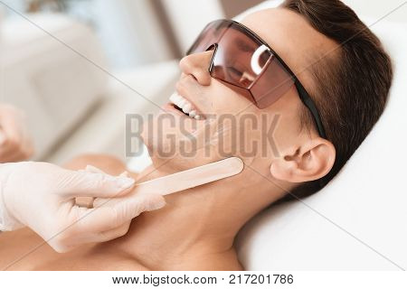 The doctor applies an epilation gel on the man's face. The man laughs, because he is ticklish. He lies on a couch in a modern beauty salon.