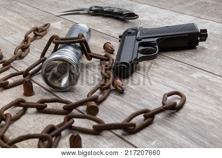 A rusty chain with a gun, a silver flashlight, bullets and a folding knife lie on a wooden background