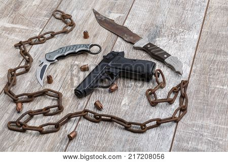 A rusty chain is a snake with a folding knife, a pistol and bullets to it and an old knife on a wooden background. Top view from an angle