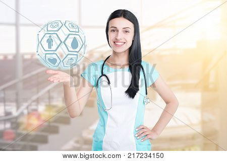 Woman doctor isolated on a clinic background, holding a graphic sphere with medical icons. Concept: medicine, innovative medical technology, healthcare.