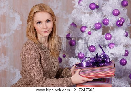 Amazing beautiful face lady girl with perfect skin and blond hairs posing for christmas holidays close to new year green pine tree and presents in studio.