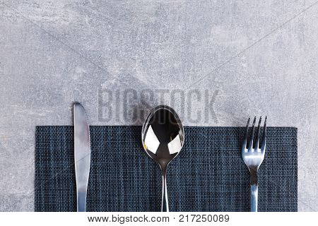 On the serving table there is a spoon, fork and knife on a dark cloth kitchen serving napkin. View from above.