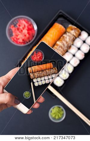 Popular Japanese food - Sushi rolls set with wasabi, ginger and chopsticks, top view. Female holding smartphone and doing picture of food.