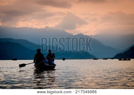 Silhouettes of people on boats at sunset on Phewa lake. Vanilla sky and mountain background.