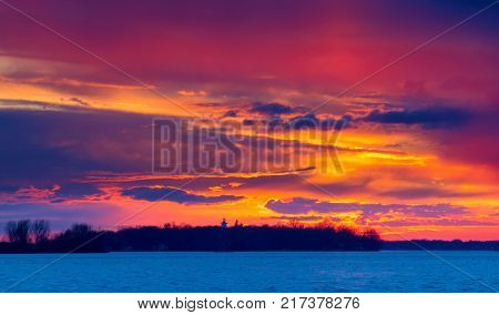 A Winter Sunset on the Lakeshore in Lachine Quebec. The silhouette of a  leafless tree, against  the blue sky and orange clouds on a cold winters afternoon, illuminate the scene.