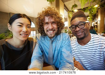 Three intercultural friends in casualwear looking at camera while gathering in cafe