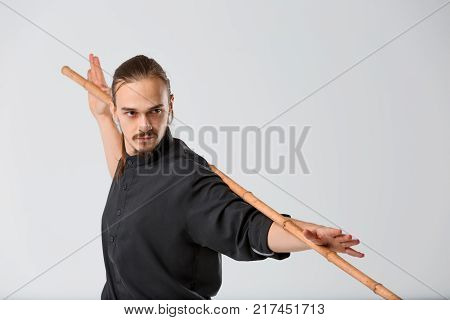 A ninja man young in a black uniform is seriously attuned. It is standing half-turned with a war stick behind the neck and holding hands close-up on a gray background
