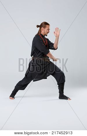 A ninja man with long hair in black uniform makes a lunging forward and is protected by his hands against a gray background. Side view close-up