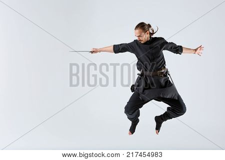 A ninja man in black uniforms jumping with a sword in his outstretched hand and bent legs on a gray background. Front view