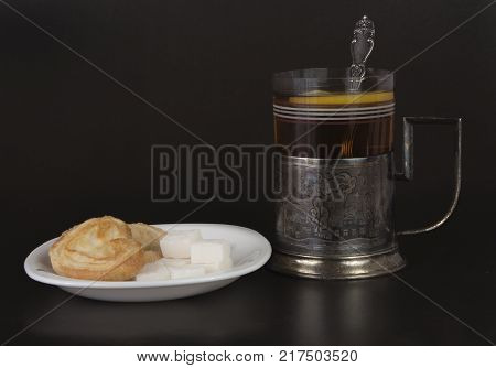 Glass with tea and teaspoon in old silver glass-holder sugar pieces and cakes on black background