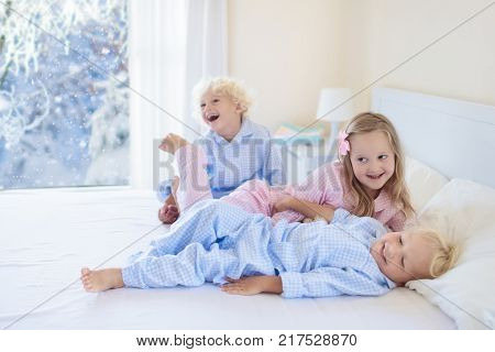 Kids playing in parents bed in winter. Children wake up in white bedroom by snow. Little boy and girl in pajamas. Christmas sleepwear and bedding for child and baby. Family morning on snowy Xmas day.
